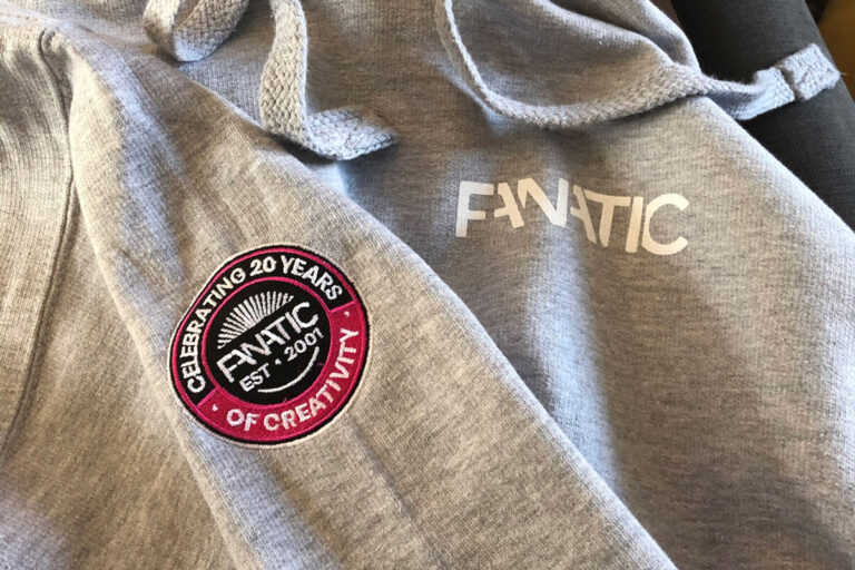 A close up of the Fanatic hoodie, which is grey and has the word 'Fanatic' printed in white on the chest, and a black and pink circular badge embroidered on the arm